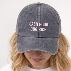 May include: A gray baseball cap with the embroidered text "CASH POOR DOG RICH" in pink. The hat has a curved brim and a classic six-panel design, suitable for casual wear.