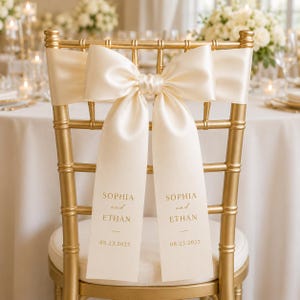 Könnte beinhalten: Ein goldfarbener Chiavari-Stuhl mit einer cremefarbenen Satinschleife. Die beiden langen, herabhängenden Enden des Bandes sind mit dem goldenen Text bedruckt: SOPHIA and ETHAN 08.23.2025. Der Stuhl verfügt über ein weißes Sitzpolster.
