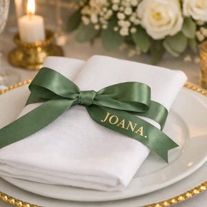 Könnte beinhalten: Eine weiße, gefaltete Serviette auf einem weißen Teller mit goldenem Rand, verziert mit einer grünen Schleife. Auf dem Band steht der Name "JOANA". Eine Kerze und Blumen sind im Hintergrund.