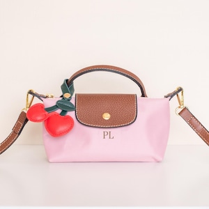 May include: A pink handbag with a brown leather handle and flap, featuring gold-tone hardware and a detachable brown strap. Adorned with a cherry charm. The initials "PL" are printed on the front.