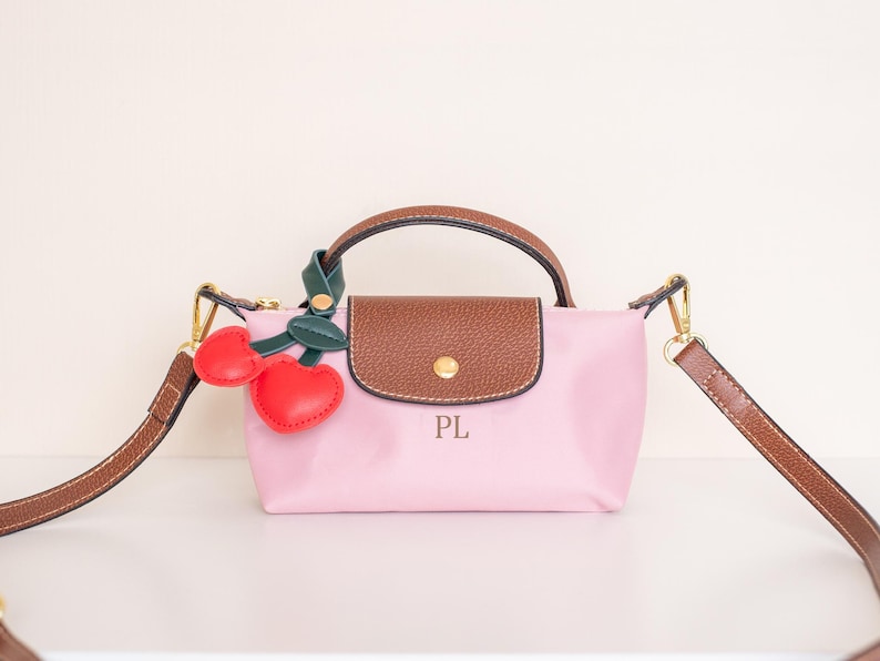 May include: A pink handbag with a brown leather handle and flap, featuring gold-tone hardware and a detachable brown strap. Adorned with a cherry charm. The initials "PL" are printed on the front.