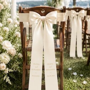 May include: A dark wood folding chair featuring a cream-coloured satin sash tied in a large bow. The long, hanging ends of the sash are printed with the text OLIVIA and BENJAMIN and the date 06.28.2025 in a simple, elegant gold-toned font.