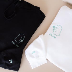 May include: Black and white hoodies with ghost graphics and text. The black hoodie has the text "Dich soor" and the white hoodie has the text "Ich mag". The sleeve of the black hoodie has the letter "B" and a heart. The sleeve of the white hoodie has the letters "J" and a heart.