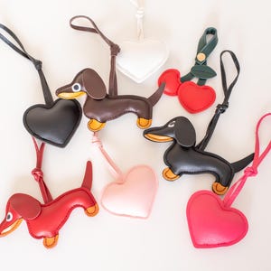 May include: A collection of leather keychains in various shapes and colors. The keychains include dachshund dogs, hearts, and cherries. The dachshunds are brown, black, and red with yellow accents. The hearts are black, white, pink, and red.