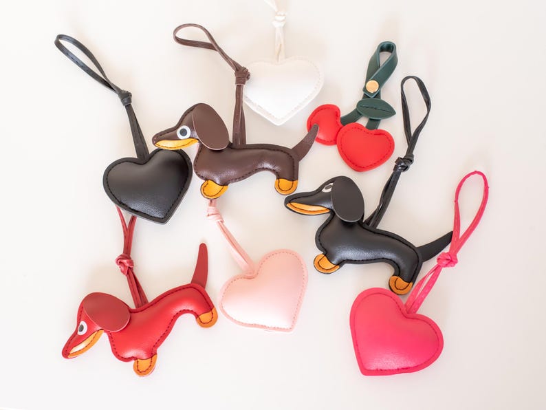 May include: A collection of leather keychains in various shapes and colors. The keychains include dachshund dogs, hearts, and cherries. The dachshunds are brown, black, and red with yellow accents. The hearts are black, white, pink, and red.