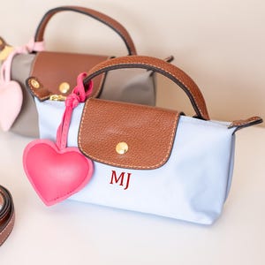 May include: Two small, rectangular handbags with brown leather handles and flaps. One is light blue, the other gray. Each has a matching heart-shaped charm. The light blue bag has the initials "MJ" in red.