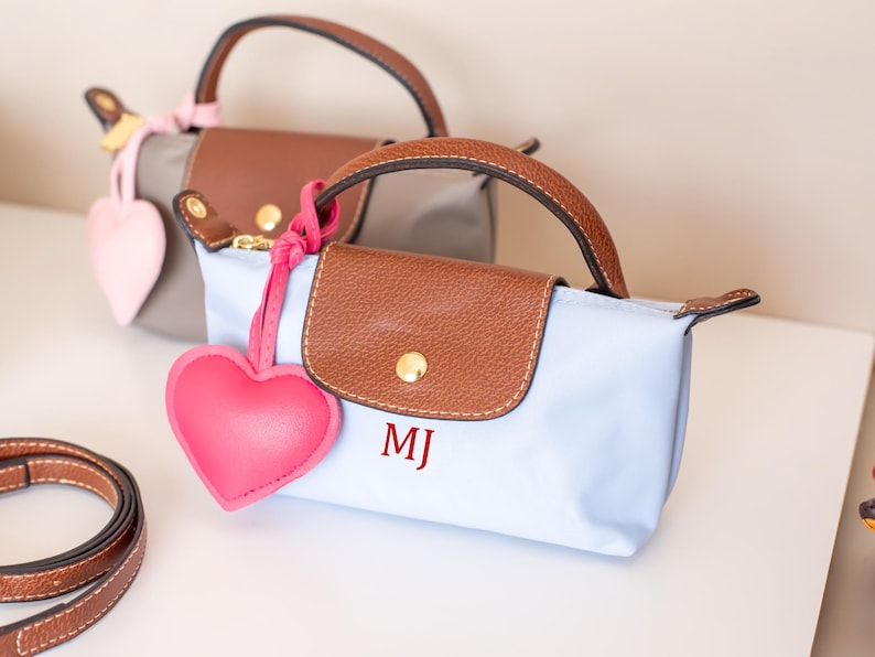 May include: Two small, rectangular handbags with brown leather handles and flaps. One is light blue, the other gray. Each has a matching heart-shaped charm. The light blue bag has the initials "MJ" in red.