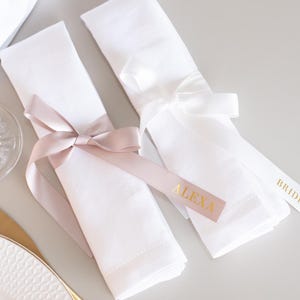 Puede incluir: Dos servilletas de lino blanco atadas con cintas de satén. Una cinta es rosa rubor con el nombre "ALEXA" en letras doradas. La otra es blanca con la palabra "BRIDE" en dorado. Decoración de mesa de boda.