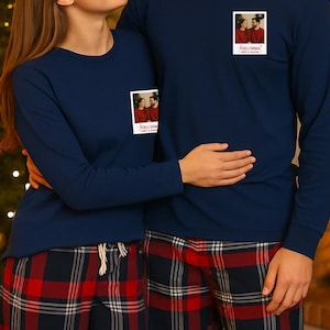 Könnte beinhalten: Passende marineblaue Langarmshirts und rot karierte Pyjamahosen. Jedes Shirt hat einen kleinen Fotodruck auf der Brust. Das Paar umarmt sich vor einem Weihnachtsbaum.