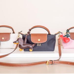 May include: Three small handbags in white, navy, and pink, each with a brown leather flap and handle. The navy bag has the initials "BA" and a dachshund charm. A brown crossbody strap is in the foreground.