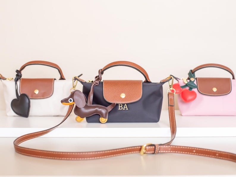 May include: Three small handbags in white, navy, and pink, each with a brown leather flap and handle. The navy bag has the initials "BA". Accessories include a heart, a dachshund, and cherries.