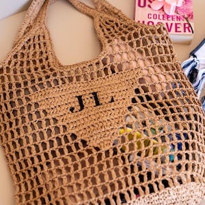Puede incluir: Un bolso tote tejido, de tejido abierto, en color beige natural. El bolso presenta las iniciales "JL" en negro en la parte delantera. El bolso está lleno de objetos coloridos. Un libro y una toalla a rayas azules y blancas están junto al bolso.