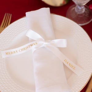 May include: A white napkin, tied with a ribbon reading "MERRY CHRISTMAS" and "DANIEL" in gold, sits on a white plate with a gold rim. Gold cutlery and a red tablecloth create a festive Christmas table setting.