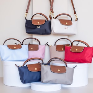 May include: A selection of small, colourful handbags with brown leather detailing. The bags are in various shades, including navy, white, light blue, pink, beige, red, and grey. Some bags have initials. Each bag has a top handle and a shoulder strap.