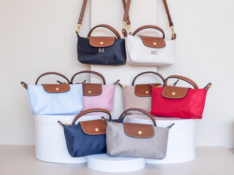 May include: Collection of small, colorful handbags with brown leather accents. The bags are in various colors, including navy, white, light blue, pink, beige, red, and gray. Some bags have initials on the front. The bags have brown leather handles and shoulder straps.