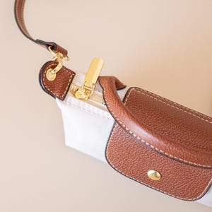 May include: Close-up of a white handbag with brown leather accents. The bag features a gold zipper and a brown leather handle. The strap is also brown leather. The bag is on a beige background.