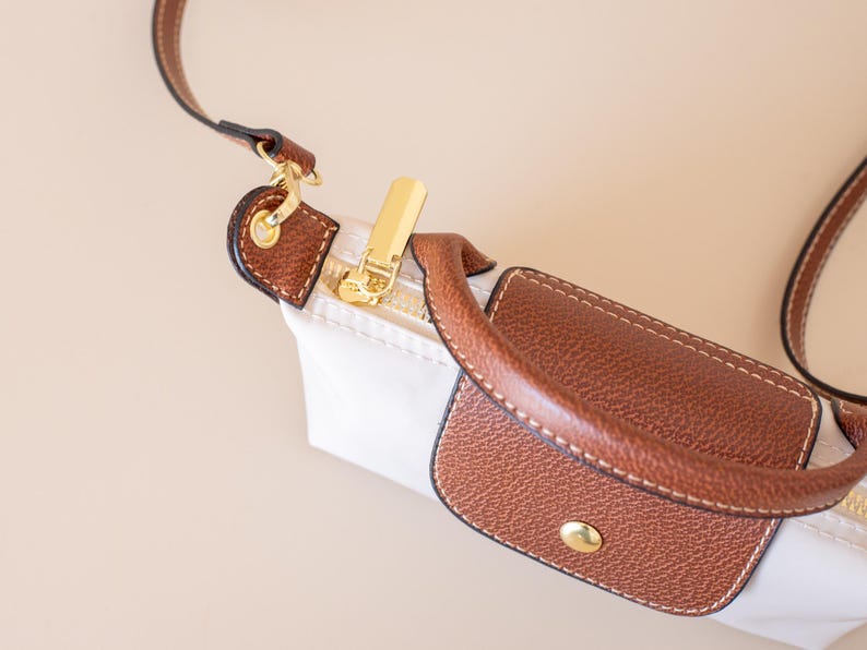 May include: Close-up of a white handbag with brown leather accents. The bag features a gold zipper and a brown leather handle. The strap is also brown leather. The bag is on a beige background.