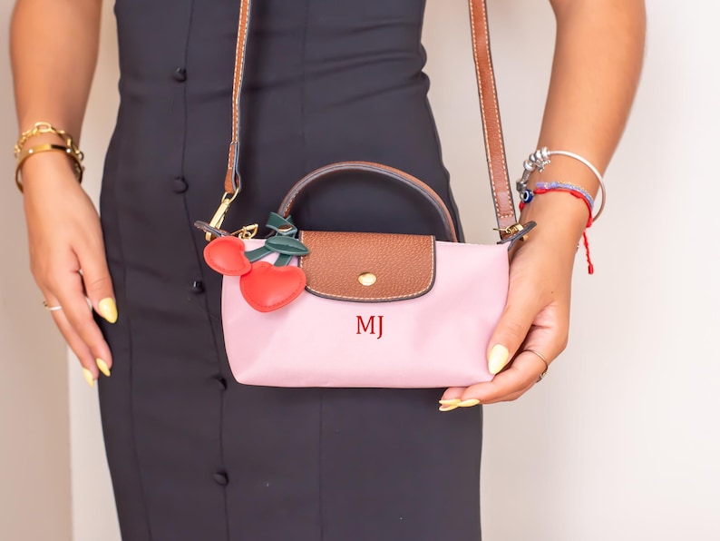 May include: A pink handbag with a brown flap and handle, featuring the initials "MJ" in red. The bag has a brown strap and a cherry charm. The person is wearing a black dress and gold bracelets.