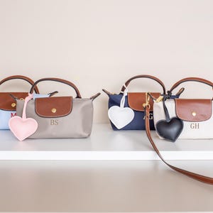 May include: Four small handbags in light blue, gray, navy, and white, each with a brown leather flap and handles. Each bag has a heart-shaped charm in pink, light pink, white, and black. The bags are personalized with initials.