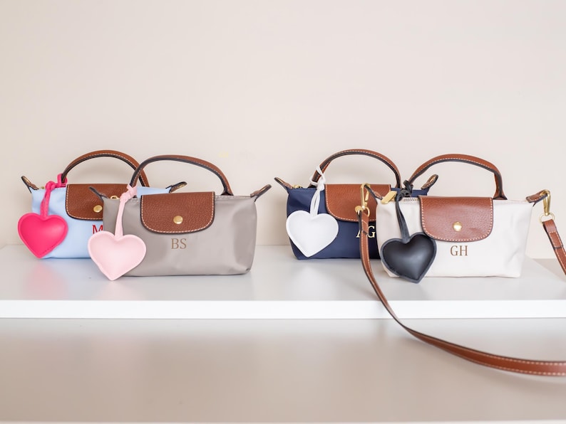 May include: Four small handbags in light blue, gray, navy, and white, each with a brown leather flap and handles. Each bag has a heart-shaped charm in pink, light pink, white, and black. The bags are personalized with initials.