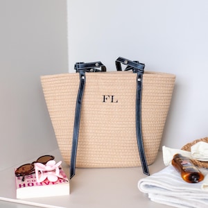 May include: A beige woven tote bag with navy blue handles and the initials "FL". Beside it are a straw hat, sunglasses, a book, sunscreen, and a folded towel. The bag is for carrying items.