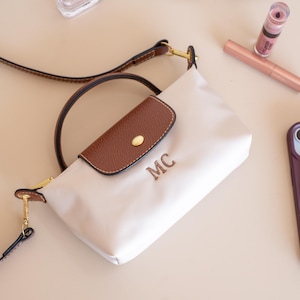 May include: Cream-colored crossbody bag with a brown flap and handle. The bag features gold-toned hardware and the initials "MC" embroidered on the front. A brown phone case, lipstick, and a tube of lip gloss are also visible.