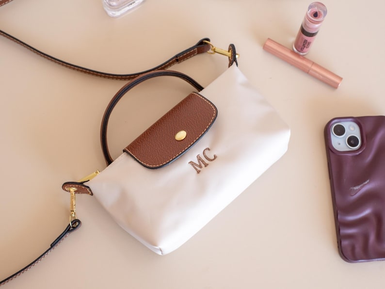 May include: Cream-colored crossbody bag with a brown flap and handle. The bag features gold-toned hardware and the initials "MC" embroidered on the front. A brown phone case, lipstick, and a tube of lip gloss are also visible.