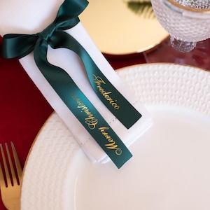 May include: A white folded napkin with a dark green ribbon tied in a bow. The ribbon has gold text that reads "Merry Christmas" and "Frederico". The napkin is on a white plate with a gold fork.