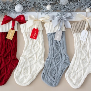 Op de afbeelding: Vier kabelgebreide kerstsokken in rood, wit, grijs en crème hangen aan een witte houten plank. Elke sok heeft een bijpassend lint en een naamlabel. Zilveren guirlande en ornamenten versieren de bovenkant.