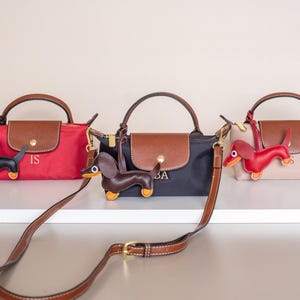 May include: Three small handbags in red, black, and beige, each with a brown flap and handle. Each bag has a dachshund charm and a brown shoulder strap. The red bag has the initials "IS", the black bag has "BA".