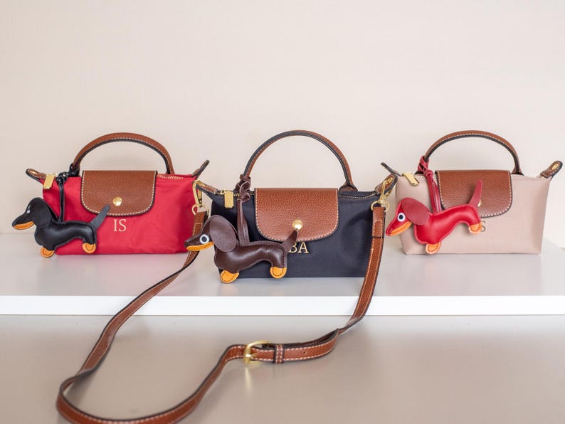 May include: Three small handbags in red, black, and beige, each with a brown flap and handle. Each bag has a dachshund charm and a brown shoulder strap. The red bag has the initials "IS", the black bag has "BA".
