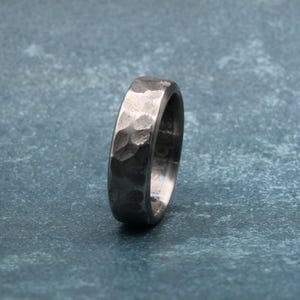 May include: A hammered silver ring with a textured surface. The ring has a band with a slightly rounded profile. The ring is set against a mottled blue-gray background.