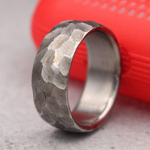 May include: A textured, hammered metal ring in grey. The ring has a wide band and a polished interior. The background includes a red object and a light grey surface.