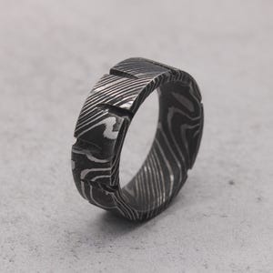 Pode incluir: Um anel escuro e padronizado com um design único. O anel apresenta uma superfície texturizada com um padrão ondulado e rodopiante. O design do anel inclui recortes retangulares. O anel é feito de metal escuro.