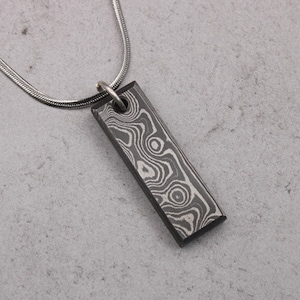 May include: A rectangular pendant necklace with a patterned design. The pendant is a dark gray color with a swirling, light gray pattern. The necklace chain is a thin, silver color. The pendant is approximately 1.5 inches long.