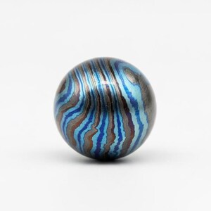 Pu&ograve; includere: Una perla rotonda e decorativa con un motivo a spirale di tonalit&agrave; blu, turchese e marrone. La perla ha una lucentezza metallica e una superficie liscia. Il design ricorda le venature del legno o un effetto marmorizzato.