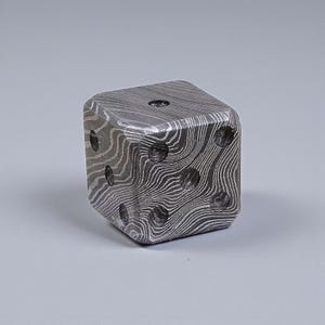 May include: A single, square die with a unique, patterned surface. The die is made of a dark metal with a swirling, wood-grain-like design. The die has six sides, each with a different number of circular indentations representing the numbers one through six.