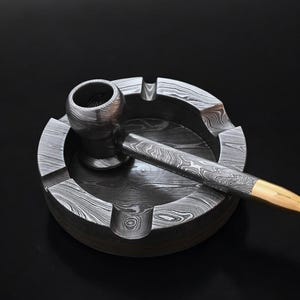 Puede incluir: Una pipa y un cenicero de acero de Damasco. La pipa tiene una boquilla dorada. El cenicero tiene ranuras para sujetar la pipa y está sobre un fondo negro.