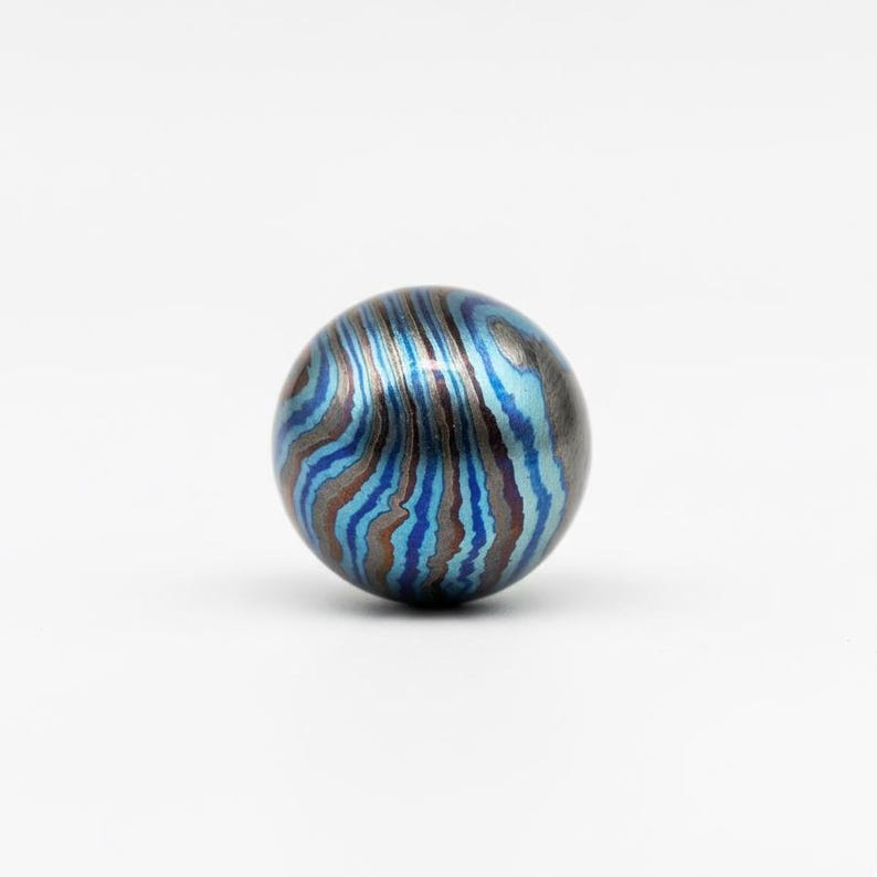 Pu&ograve; includere: Una perla rotonda e decorativa con un motivo a spirale di tonalit&agrave; blu, turchese e marrone. La perla ha una lucentezza metallica e una superficie liscia. Il design ricorda le venature del legno o un effetto marmorizzato.