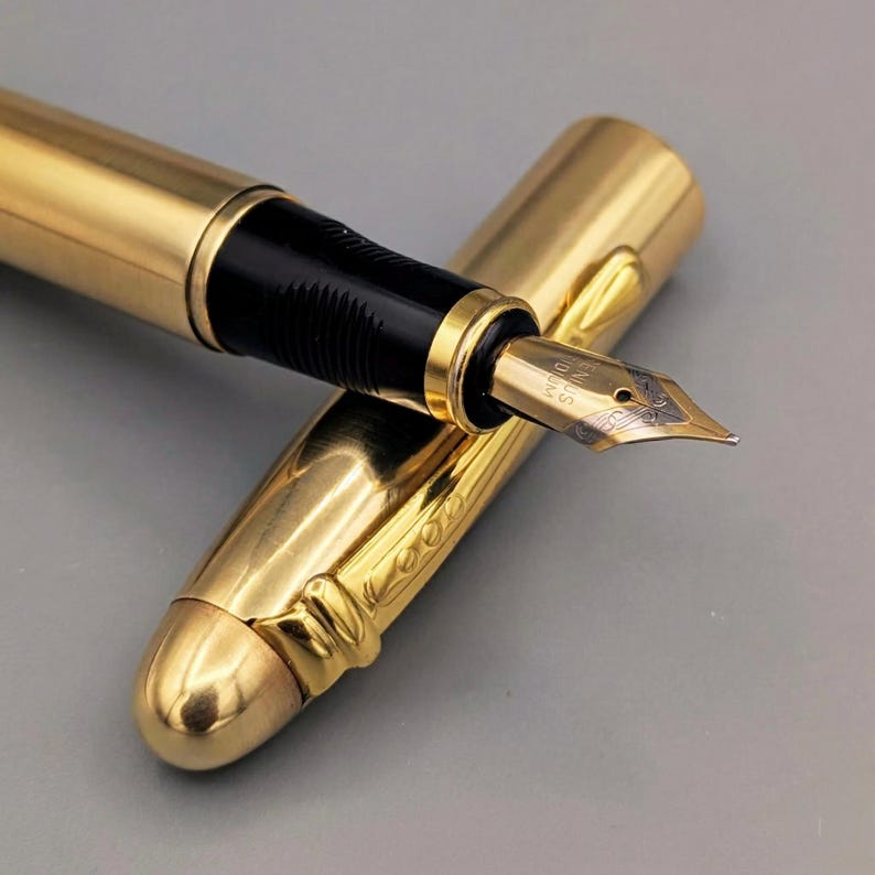 May include: A gold-colored fountain pen with a black grip and a gold nib. The pen's cap is off, revealing the nib and the pen's design. The pen is lying on a gray surface.