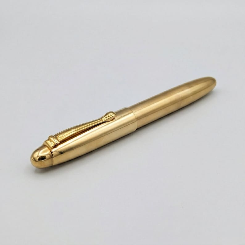 May include: A gold-colored pen with a sleek, streamlined design. The pen features a clip and a rounded tip. The pen is made of a shiny, metallic material and is set against a plain background. The pen is a writing instrument.