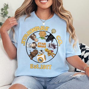 May include: Light blue Star Wars Co. t-shirt featuring a circular design with various Star Wars characters, including BB-8, Chewbacca, and Yoda. The design also includes the words 'Star Wars' and 'Est. 1977' in yellow lettering. The shirt is made of a soft material.