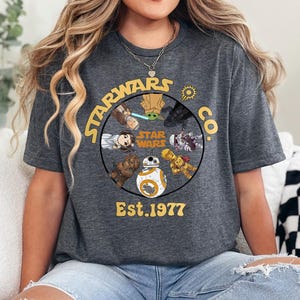 Peut inclure: T-shirt gris fonc&eacute; avec un motif circulaire repr&eacute;sentant des personnages de Star Wars et le texte "STARWARS CO. Est. 1977". Le design comprend des personnages comme Yoda et BB-8 de la franchise Star Wars.