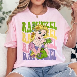 May include: Light pink t-shirt featuring a graphic of Rapunzel with the word "Rapunzel" in a retro font. The design includes a cartoon illustration of Rapunzel holding a green chameleon. The text "LIGHT PINK" is at the bottom.