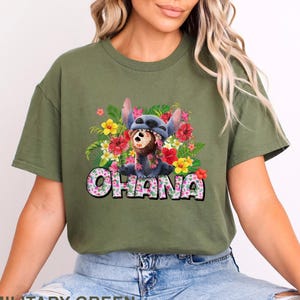 May include: Military green t-shirt featuring a colorful graphic of Stitch wearing a floral lei and a Stitch hat, surrounded by flowers. The word "Ohana" is printed in a floral font.
