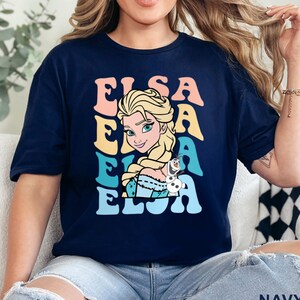 May include: Navy blue t-shirt featuring a colorful Elsa graphic. The design includes the name 'ELSA' stacked vertically in pastel colors, with an illustration of Elsa from Frozen. The character is blonde with a braid, and a small Olaf figure is also present.