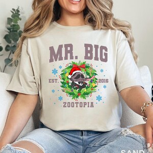 以下が含まれることがあります： 砂色のTシャツ。テキスト「MR. BIG」の下に、サンタ帽とタキシードを着たアライグマのグラフィックがあり、クリスマスのリースで囲まれています。「EST. 2016」と「ZOOTOPIA」の文字も表示されています。