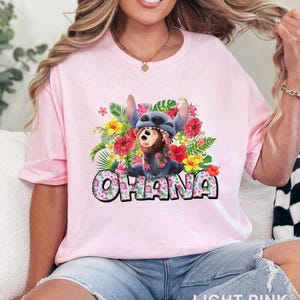 May include: Light pink t-shirt with a colorful graphic of Stitch wearing a Hawaiian lei and a hat. The word 'OHANA' is printed in a floral font below the character. The shirt is made of soft fabric and has a crew neck. The overall theme is Hawaiian and Disney.