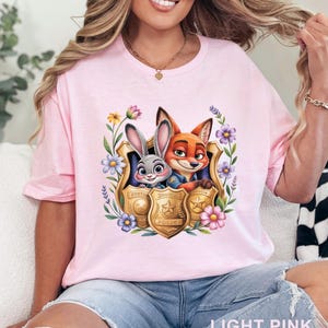 Puede incluir: Camiseta rosa claro con un gr&aacute;fico colorido de Judy Hopps y Nick Wilde de Zootopia dentro de una placa de polic&iacute;a, rodeada de flores. El texto "LIGHT PINK" est&aacute; en la parte inferior. Cuello redondo.