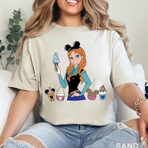 May include: Sand-colored t-shirt featuring a cartoon illustration of a character with red hair and a blue ice cream cone. The character is surrounded by ice cream treats. The word "SAND" is printed at the bottom.